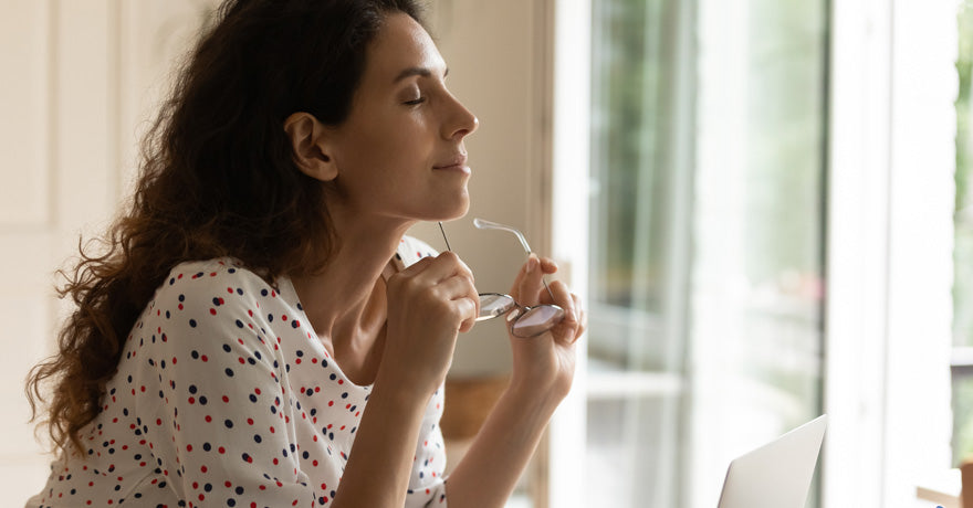 Woman holding glasses, eye closed, at window