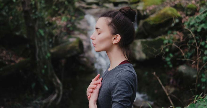 Woman with eyes closed and hands on heart in front of waterfall